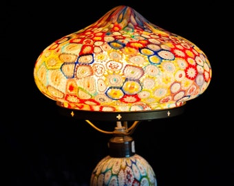 May include: A colorful, vintage table lamp with a patterned glass shade and base. The shade is a vibrant mix of red, yellow, blue, and white, while the base has a similar design. The lamp is illuminated, casting a warm glow.