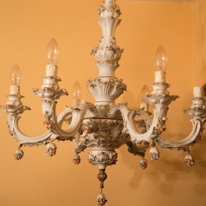 May include: An ornate white porcelain chandelier with six arms, adorned with floral accents in pink, orange, and green. The fixture has a decorative central column and is suspended from the ceiling.