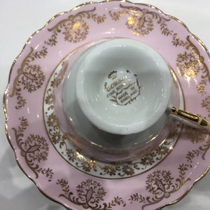 May include: A pink teacup and saucer set with gold trim and floral designs. The saucer has a scalloped edge. The teacup has the words "Coronation Fine Bone China Made in England" on the bottom.