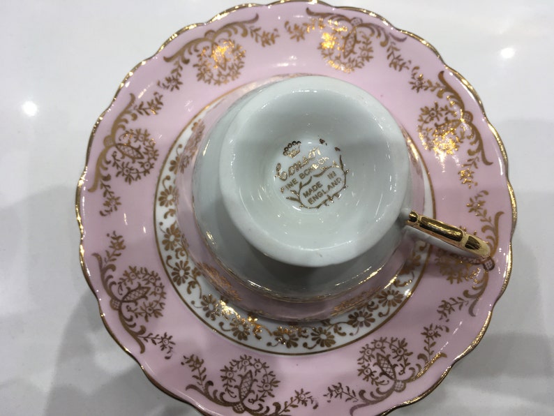 May include: A pink teacup and saucer set with gold trim and floral designs. The saucer has a scalloped edge. The teacup has the words "Coronation Fine Bone China Made in England" on the bottom.
