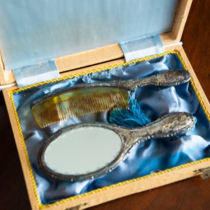 Op de afbeelding: Een vintage toilettasje met een zilveren handspiegel en een kam in een houten doos bekleed met blauw fluweel.