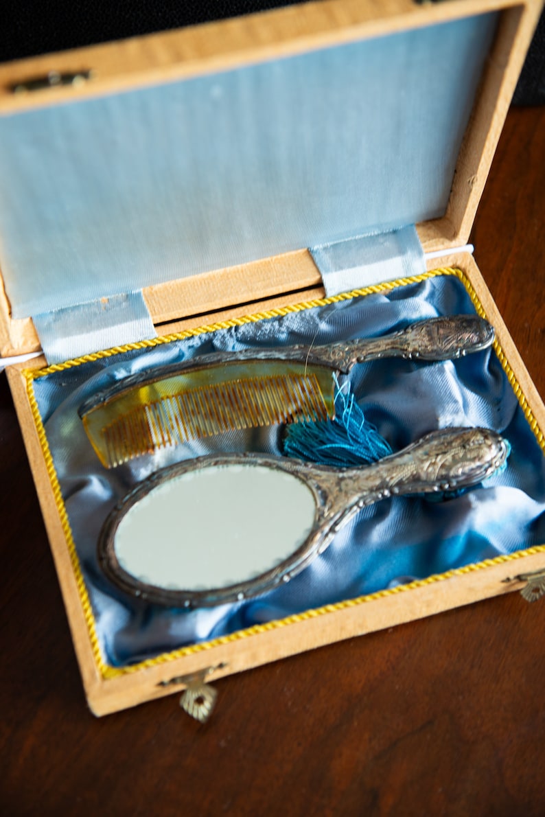Op de afbeelding: Een vintage toilettasje met een zilveren handspiegel en een kam in een houten doos bekleed met blauw fluweel.