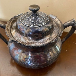 May include: A silver sugar bowl with a lid. The lid has an ornate design and the bowl has a scalloped edge.