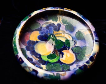 May include: A round ceramic dish featuring a colourful design of fruit and leaves. The dish is painted in shades of blue, green, yellow, and purple.