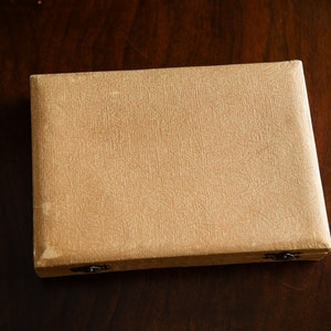 Op de afbeelding: Een beige, rechthoekige doos met twee kleine, metalen sluitingen. De doos heeft een getextureerd oppervlak en is gemaakt van karton.