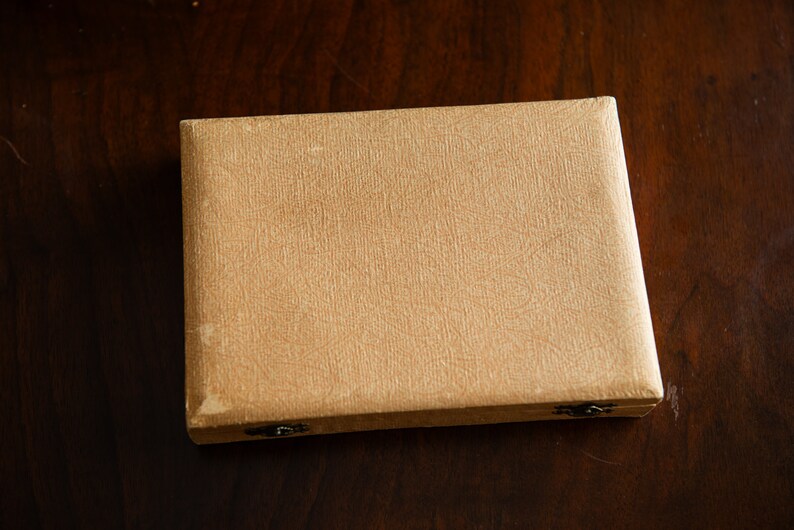 Op de afbeelding: Een beige, rechthoekige doos met twee kleine, metalen sluitingen. De doos heeft een getextureerd oppervlak en is gemaakt van karton.