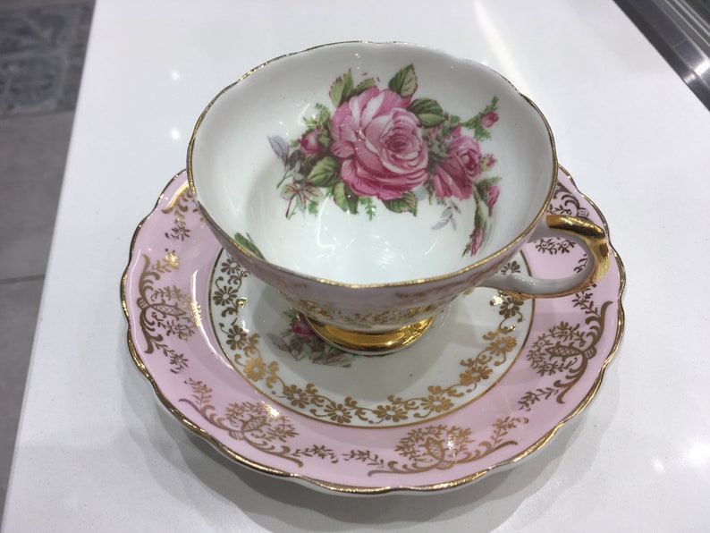 May include: A pink teacup and saucer set with a floral design. The cup and saucer are decorated with gold trim and a pink rose pattern.