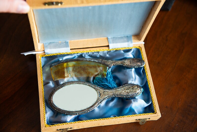 Op de afbeelding: Een houten doos met een blauwe satijnen voering, die een handspiegel met een zilveren handvat en een kam met een schildpadschelp handvat bevat.