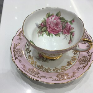 May include: A pink and gold teacup and saucer set with a floral design. The cup features a large pink rose with green leaves and smaller pink roses. The saucer has a pink rim with a gold floral pattern.