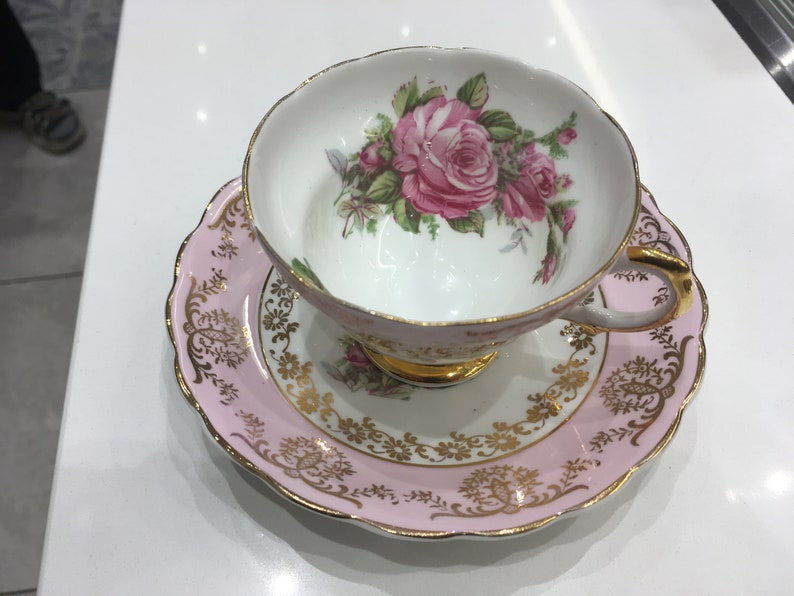 May include: A pink and gold teacup and saucer set with a floral design. The cup features a large pink rose with green leaves and smaller pink roses. The saucer has a pink rim with a gold floral pattern.