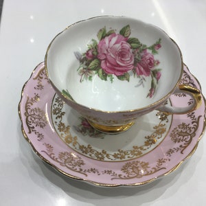 May include: A pink teacup and saucer set with a gold trim and floral design. The teacup features a large pink rose with green leaves.
