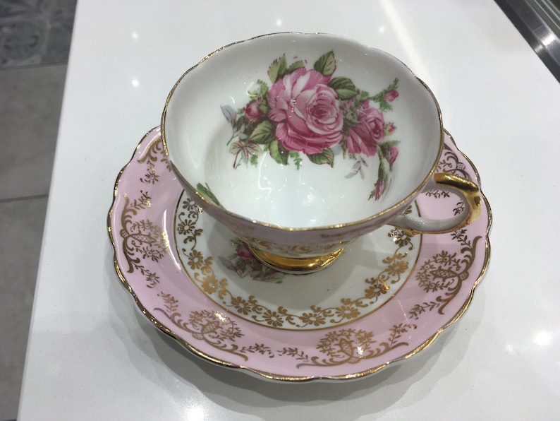 May include: A pink teacup and saucer set with a gold trim and floral design. The teacup features a large pink rose with green leaves.