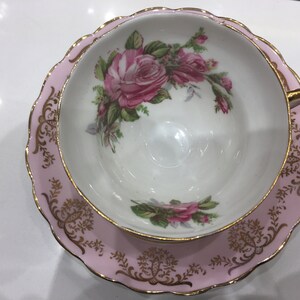 May include: A pink teacup and saucer set with a gold trim and floral design. The cup and saucer feature a delicate floral pattern with pink roses and green leaves.