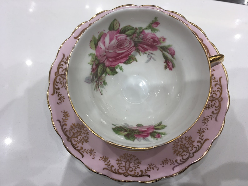 May include: A pink teacup and saucer set with a gold trim and floral design. The cup and saucer feature a delicate floral pattern with pink roses and green leaves.