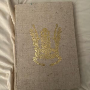 May include: A hardcover book with a beige linen cover. A gold floral design is centered on the front. The book is lying on a white, textured surface.
