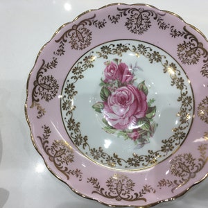 May include: A pink and gold saucer with a floral design featuring pink roses and gold accents. The saucer has a scalloped edge.