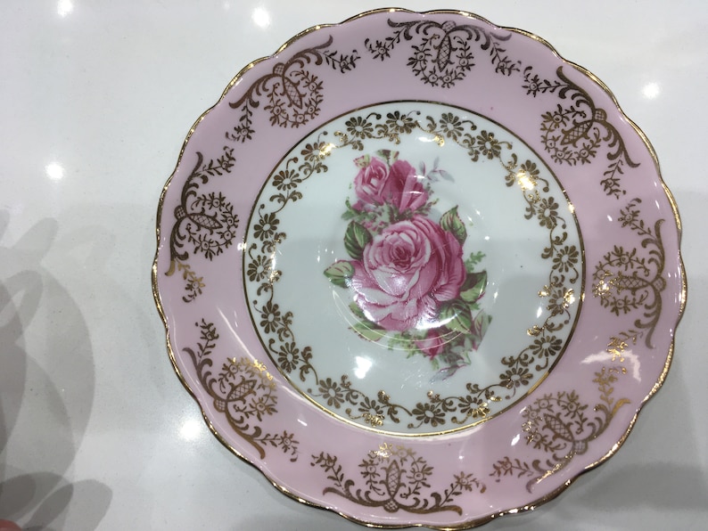 May include: A pink and gold saucer with a floral design featuring pink roses and gold accents. The saucer has a scalloped edge.