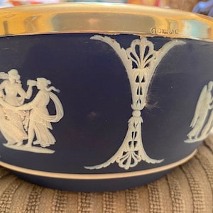 May include: A blue and white ceramic bowl with a gold rim. The bowl features a white cameo design of figures in a classical setting. The bowl is marked "Wedgwood" on the bottom.