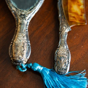 Op de afbeelding: Een vintage zilveren handspiegel en kamset met sierlijke details. De spiegel heeft een blauwe kwast aan de handgreep.