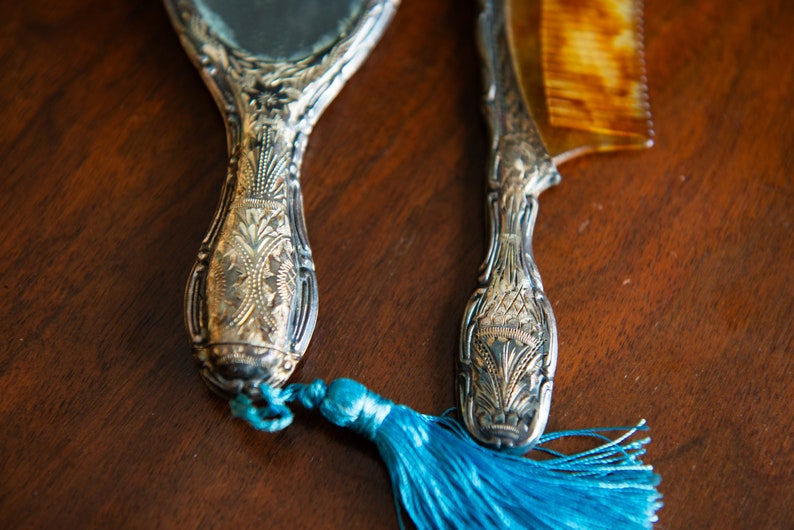 Op de afbeelding: Een vintage zilveren handspiegel en kamset met sierlijke details. De spiegel heeft een blauwe kwast aan de handgreep.