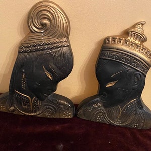 May include: A pair of black and gold ceramic wall sculptures depicting stylised Asian figures. The figures are wearing elaborate headdresses and ornate clothing.