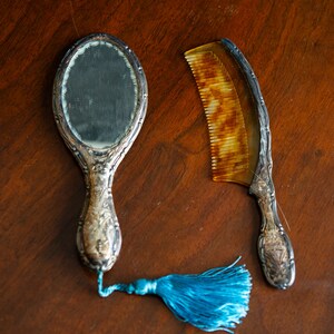 Op de afbeelding: Een verzilverde handspiegel met een blauwe kwast en een bijpassende verzilverde kam met een bruin schildpaden handvat.