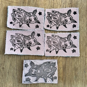 May include: Five fabric patches and a stamp featuring a stylized moth design. The patches are rectangular with a pink background, black moth illustration, and black star accents. The stamp is a gray and white block with the same moth design.