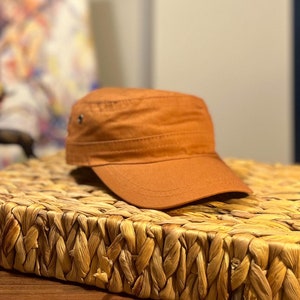 Könnte beinhalten: Eine braune Canvas-Kappe mit einem kurzen Schirm und einem kleinen Knopf oben. Die Kappe liegt auf einer geflochtenen Strohmatte.