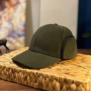 Puede incluir: Una gorra de béisbol verde oscuro con visera negra y un ribete negro alrededor de las orejeras. La gorra está sobre una estera de paja tejida.