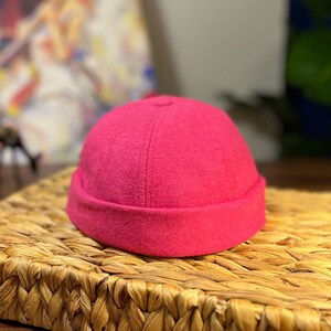 以下が含まれることがあります： 折り畳まれた縁のある明るいピンクのウール製のビーニー帽。帽子は、編まれたわらマットの上に置かれています。