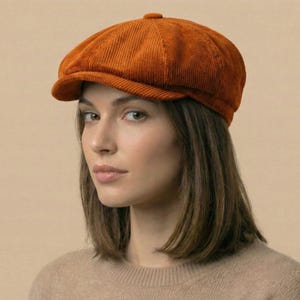 May include: An orange corduroy newsboy cap. The cap features a rounded crown and a short, stiff peak. The fabric has a ribbed texture. The cap is a warm, earthy tone, suitable for autumn or winter wear. A classic style.