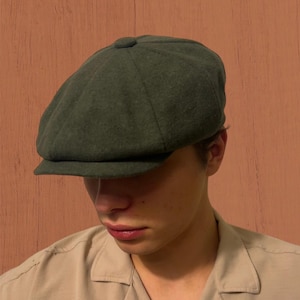Puede incluir: Gorra de estilo newsboy en verde oscuro, con una corona redondeada y una visera corta y rígida. La gorra está hecha de una tela texturizada y se lleva baja sobre la frente. Un estilo clásico, adecuado para varios conjuntos.