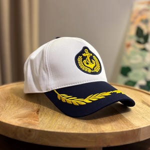 Könnte beinhalten: Weiß-marineblaue Baseballkappe mit nautischem Design. Die Kappe hat ein goldenes Ankeremblem und ein goldenes Lorbeerblatt-Design auf dem Schirm. Die Kappe liegt auf einem Holztablett.