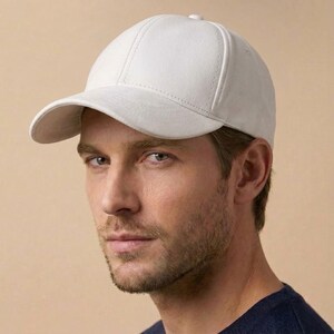 May include: A solid white baseball cap featuring a structured six-panel crown, a curved brim, and visible tonal stitching along the seams. The cap has a smooth, matte finish and a button detail at the top center of the crown, providing a clean look.