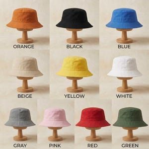 Puede incluir: Una colección de once sombreros de pescador exhibidos en soportes de madera, cada uno etiquetado con su color: naranja, negro, azul, beige, amarillo, blanco, gris, rosa, rojo y verde. Estos sombreros lisos tienen un ala inclinada y copa plana.