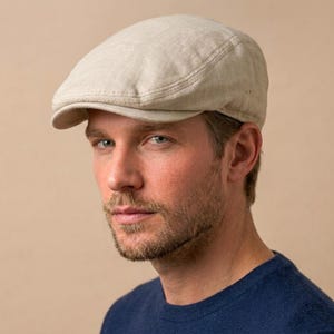 Op de afbeelding: Een beige platte pet met een ronde kroon en een korte, stijve klep. De pet heeft een constructie van gestructureerde stof met zichtbare stiksels langs de naden en een gestructureerd paneelontwerp dat laag op het voorhoofd rust.