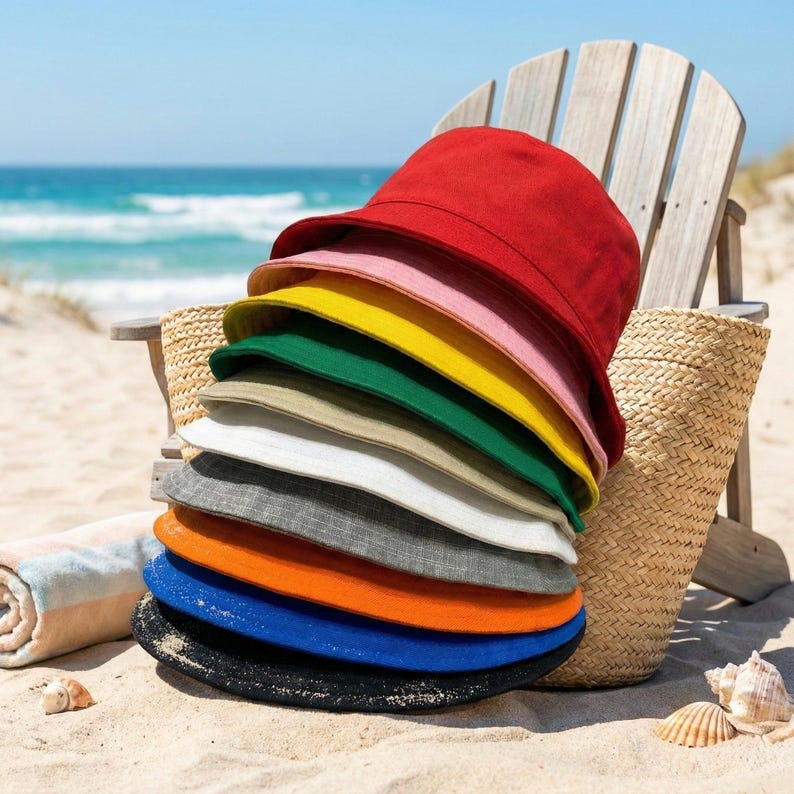 May include: A stack of colorful bucket hats in various shades, including red, pink, yellow, green, and blue, are displayed on a sandy beach. A wooden chair and a woven tote bag are in the background, with the ocean visible.