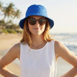 May include: A woman wearing a bright blue bucket hat and tortoiseshell sunglasses. She is also wearing a white sleeveless top and a delicate gold necklace. The background shows a beach with palm trees and the ocean.