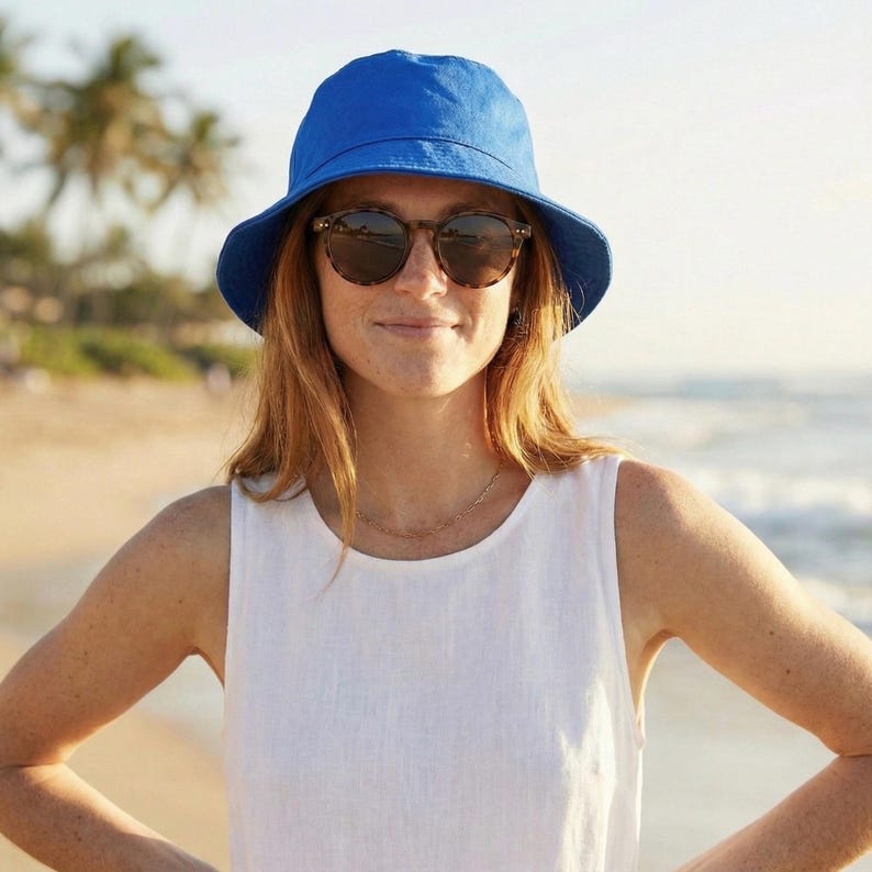 May include: A woman wearing a bright blue bucket hat and tortoiseshell sunglasses. She is also wearing a white sleeveless top and a delicate gold necklace. The background shows a beach with palm trees and the ocean.