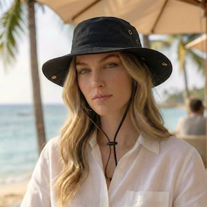 Puede incluir: Un sombrero de pescador negro con ala ancha y ojales. El sombrero tiene una correa para la barbilla y lo lleva una persona que lleva una camisa blanca abotonada. El fondo incluye una escena de playa con palmeras y una sombrilla.