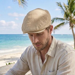 May include: A tan-colored, flat cap with a curved brim. The cap is made of a textured fabric and has a stitched seam detail. The image shows the cap being worn outdoors, with a beach and ocean in the background. The cap is part of a casual wear collection.