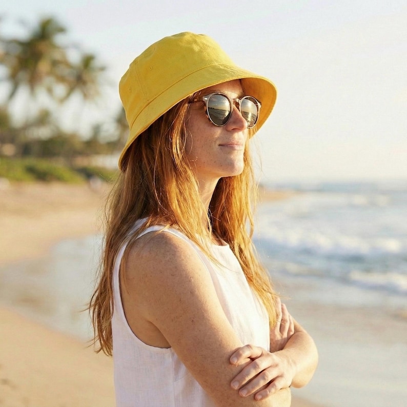 May include: A person wearing a yellow bucket hat and sunglasses, standing on a beach. The hat is a bright yellow color, and the sunglasses have a tortoiseshell pattern. The person is wearing a white sleeveless top.