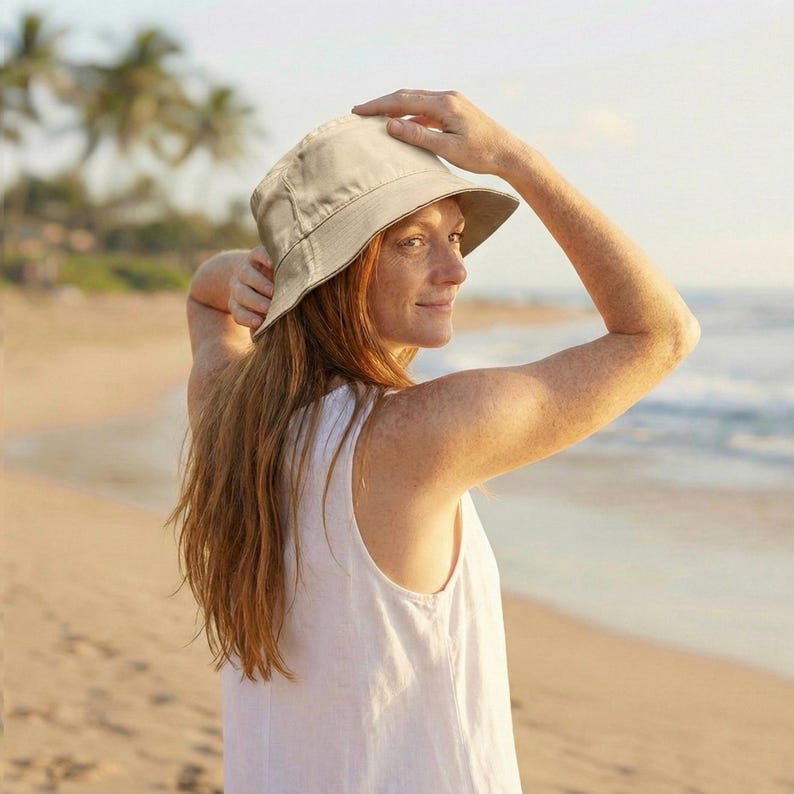 May include: A beige bucket hat with a wide brim. The hat is made of a soft fabric and is worn by a person on a beach. The person is wearing a white sleeveless top. The background includes the ocean and palm trees.