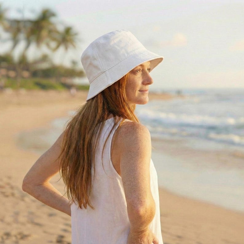 May include: A white bucket hat with a slightly angled brim. The hat is made of a soft, light-colored fabric. The hat is worn by a person on a beach with the ocean in the background.