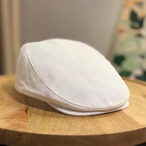 May include: A solid white flat cap featuring a structured crown and a short, stiff brim. The fabric displays a subtle, fine-textured weave pattern throughout the surface of the cap, which maintains a rounded, low-profile shape.