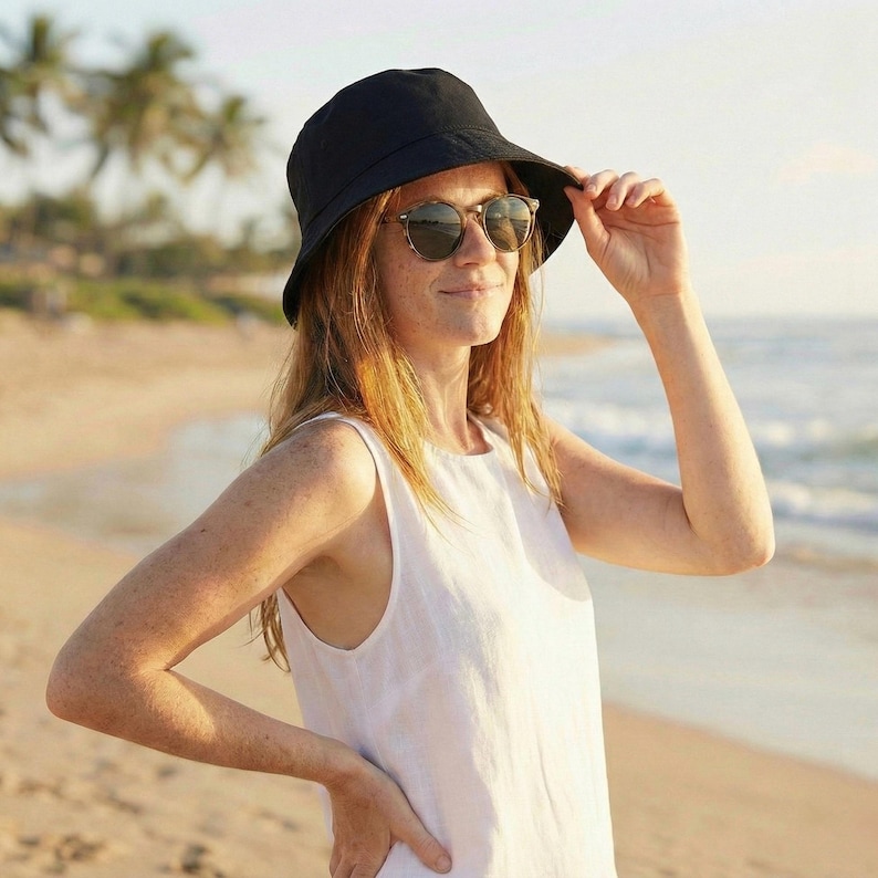 May include: A woman wearing a black bucket hat and sunglasses on a sunny beach. The hat is covering the top of her head, and the sunglasses are round with a tortoiseshell pattern. She is wearing a white sleeveless top.