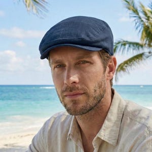 Op de afbeelding: Een marineblauwe pet van een textuurstof. De pet heeft een ronde kroon en een korte, stijve klep. De achtergrond toont een strand met palmbomen en de zee.