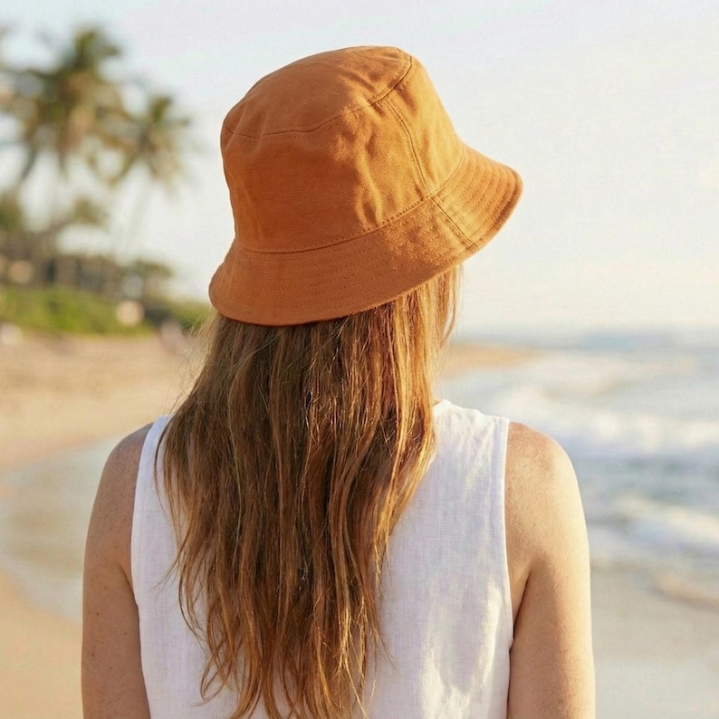 May include: A person wearing a burnt orange bucket hat and a white sleeveless top, standing on a beach. The hat is made of a textured fabric and has a wide brim. The person's long, wavy hair is visible.