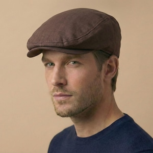 Könnte beinhalten: Eine braune Flatcap aus strukturiertem Stoff. Die Kappe hat einen kurzen Schirm und eine runde Krone. Die Person trägt einen blauen Pullover. Der Hintergrund ist einfarbig beige.