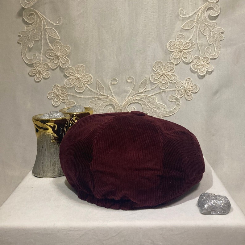 Peut inclure: Une casquette de newsboy en velours c&ocirc;tel&eacute; bordeaux avec une couronne arrondie et une visi&egrave;re plate.